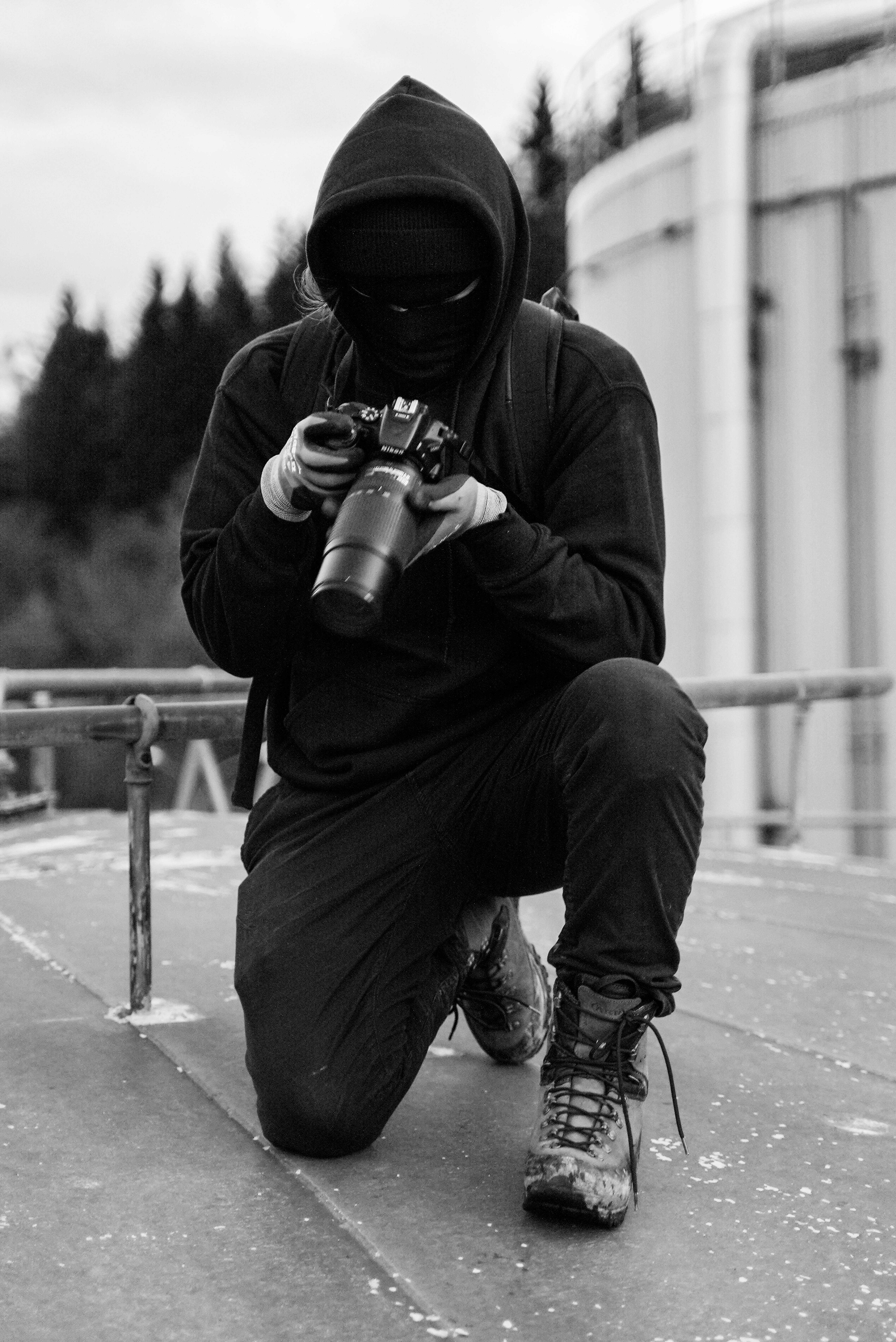 Black and white image of a hooded photographer kneeling with a camera outdoors.