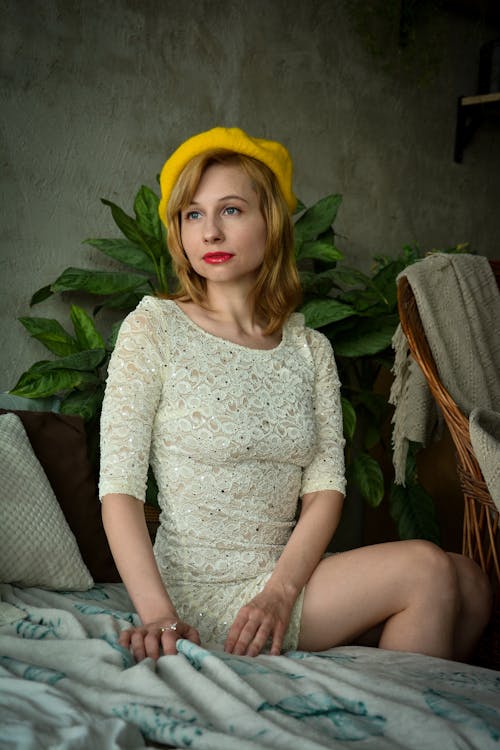 Charming young female in vintage dress and yellow beret sitting on comfortable sofa in studio in front of green plant near armchair