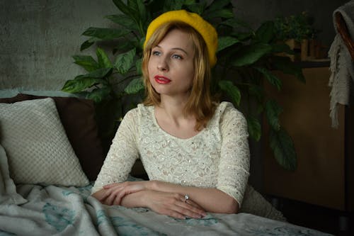 Thoughtful young female in retro style dress and yellow hat leaning on bed and looking away while posing in cozy interior studio in natural daylight