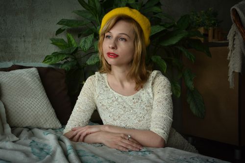 Free Thoughtful young female in retro style dress and yellow hat leaning on bed and looking away while posing in cozy interior studio in natural daylight Stock Photo