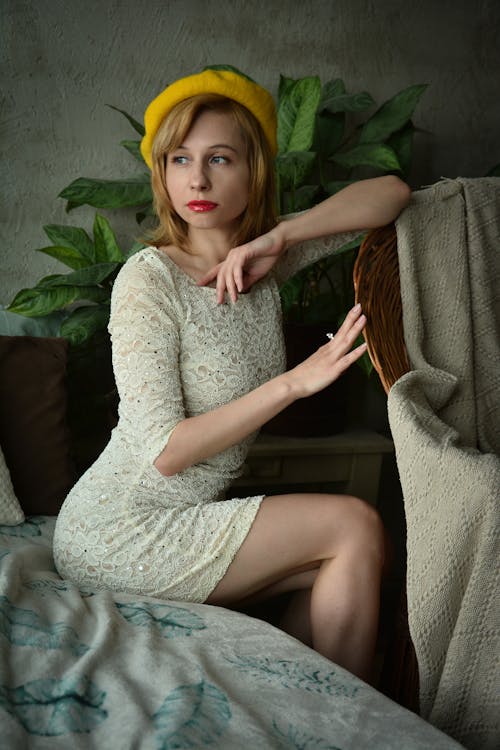 Free Young attractive blond lady wearing retro clothes sitting on edge of bed and leaning arm on plaited chair while looking away Stock Photo
