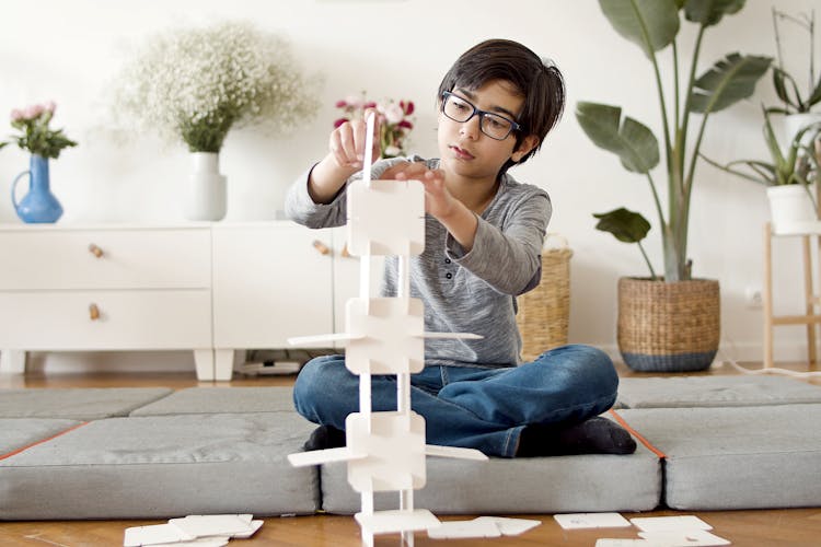 A Young Boy In Gray Long Sleeves Sitting While Building A Paper Tower