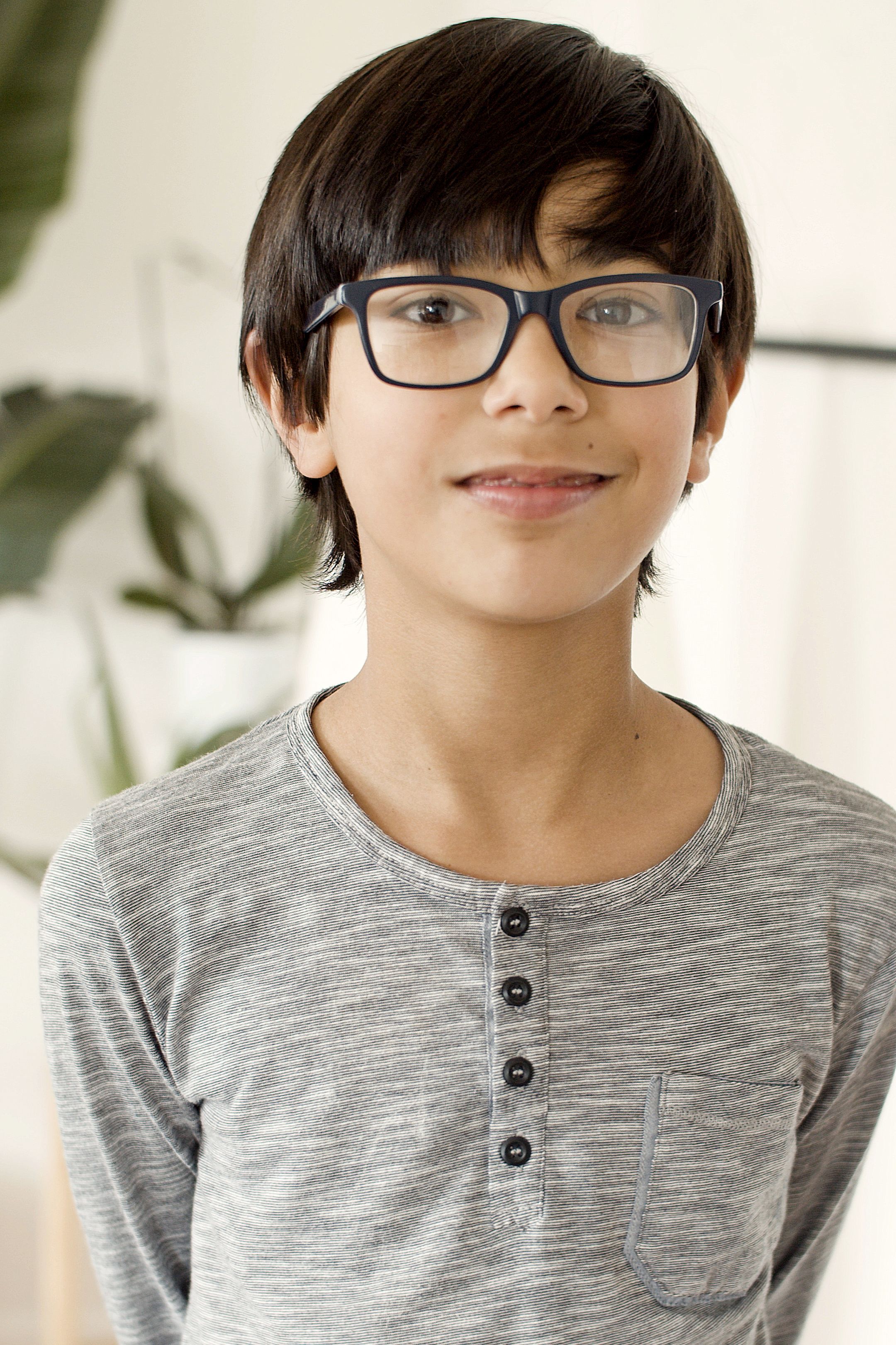 Portrait of a young boy wearing glasses and a gray shirt, smiling indoors.