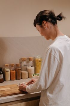 A woman chopping ingredients in a cozy kitchen setup with glass jars on the counter.