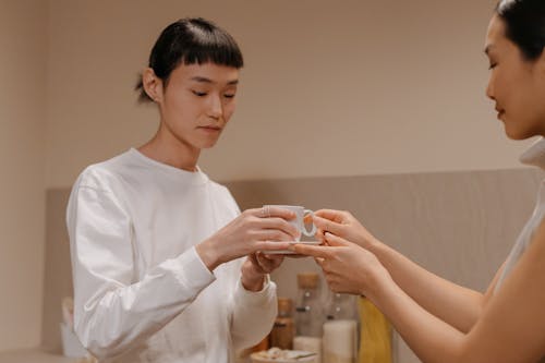 Side view of crop adult Asian woman giving cup of freshly brewed coffee to teenage daughter while standing together in kitchen during breakfast