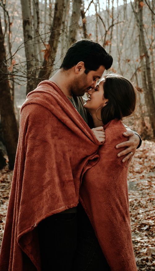 Free Side view of Hispanic male hugging and kissing smiling female while standing and covering with warm blanket in autumn park in daylight Stock Photo