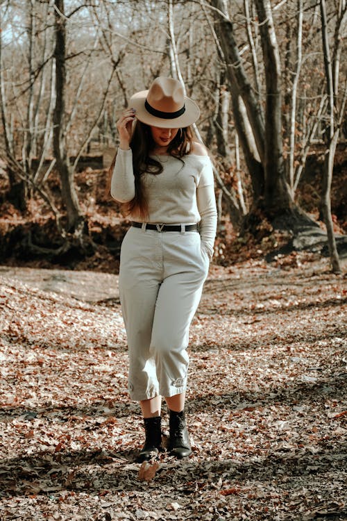Full body of unrecognizable female model wearing trendy outfit standing and hiding behind hat while walking in park in fall in daylight