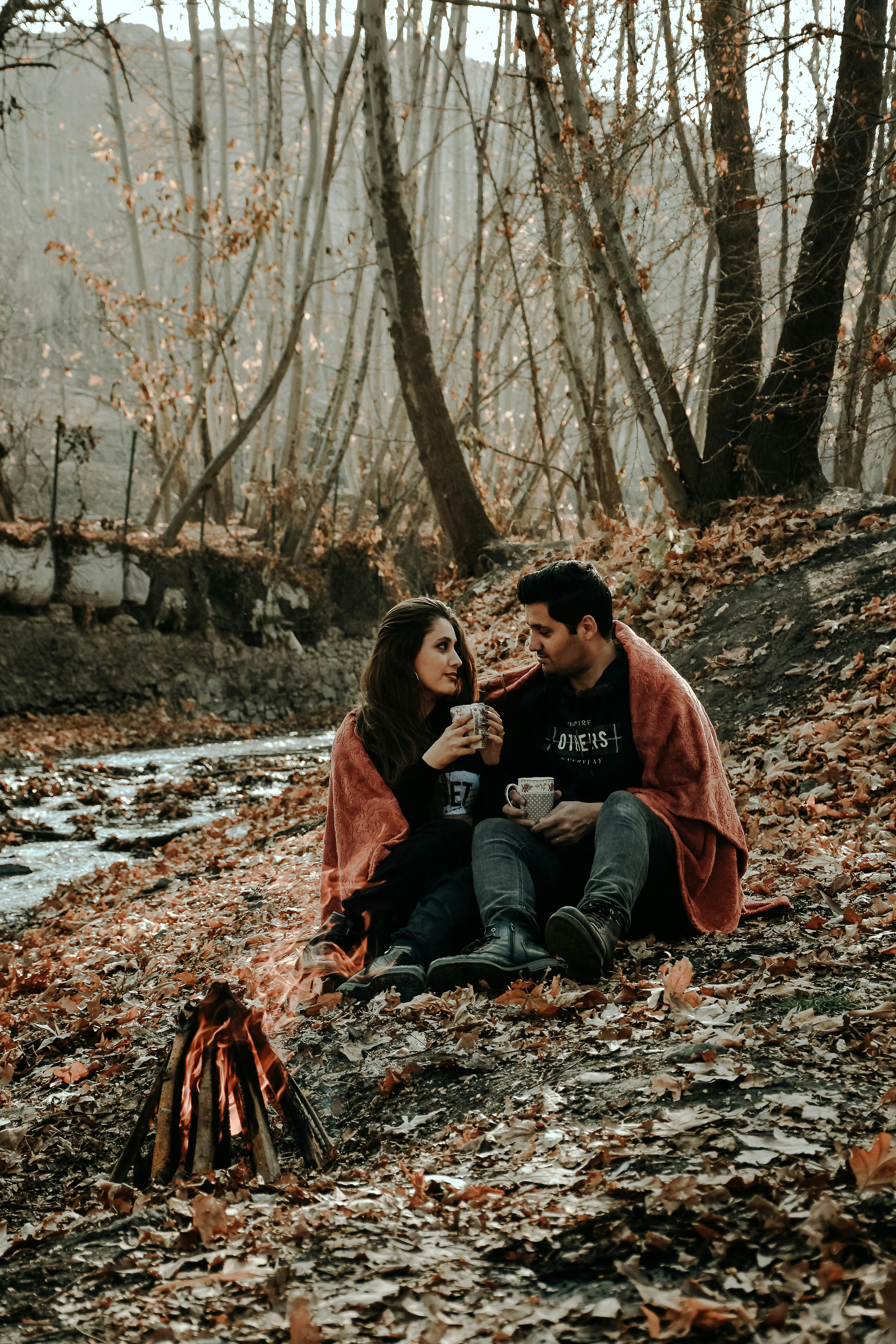 Full body of happy Hispanic couple wearing warm clothes drinking hot coffee and looking at each other while sitting near bonfire in fall