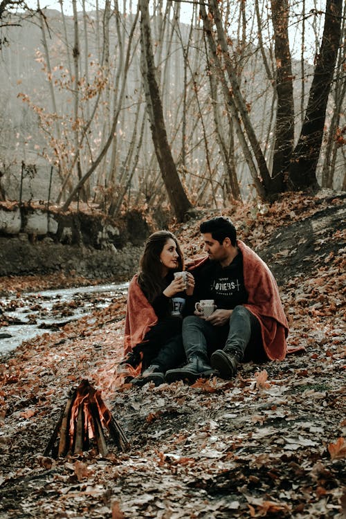 Full body of happy Hispanic couple wearing warm clothes drinking hot coffee and looking at each other while sitting near bonfire in fall