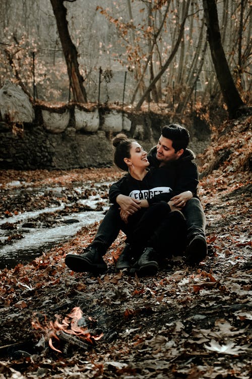 Full body of Hispanic couple in cozy outfits laughing and looking at each other while hugging near bonfire in park in daylight