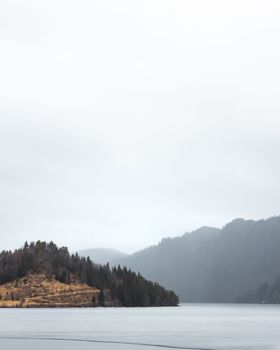 A picturesque view of a misty lake surrounded by hills under an overcast sky, conveying tranquility.