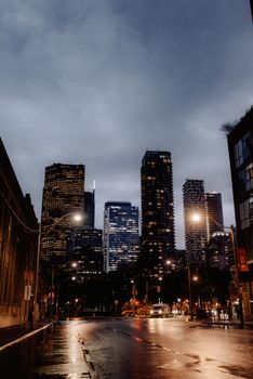 From low facades of modern illuminated skyscrapers and wet asphalt road against cloudy sky on rainy evening in city