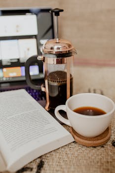A warm, inviting setup with a French press, cup of coffee, and an open book on a table.