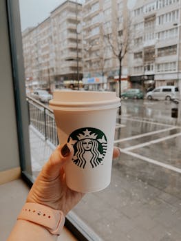 Woman holding a paper coffee cup inside a café with a rainy city view outside.