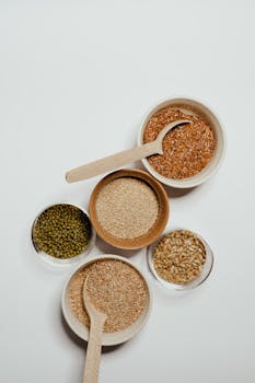 Top view of assorted grains in bowls with wooden spoons on a minimalist background.