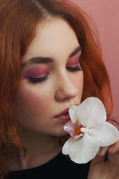 Serious young lady in colorful makeup with white orchid flower with closed eyes on pink background in light studio