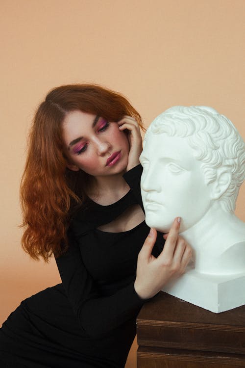 Thoughtful young female in black dress with makeup near white man sculpture on stand on beige background in light room