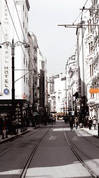 Busy city street with tram tracks lined with buildings under a bright sky.