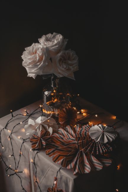 Heap of colorful paper decorations placed on table with glowing lights and vase with blooming flowers in room on black background