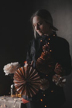 Sad skilled female in black outfit with creative handmade paper decorations and glowing garland standing near table on dark background
