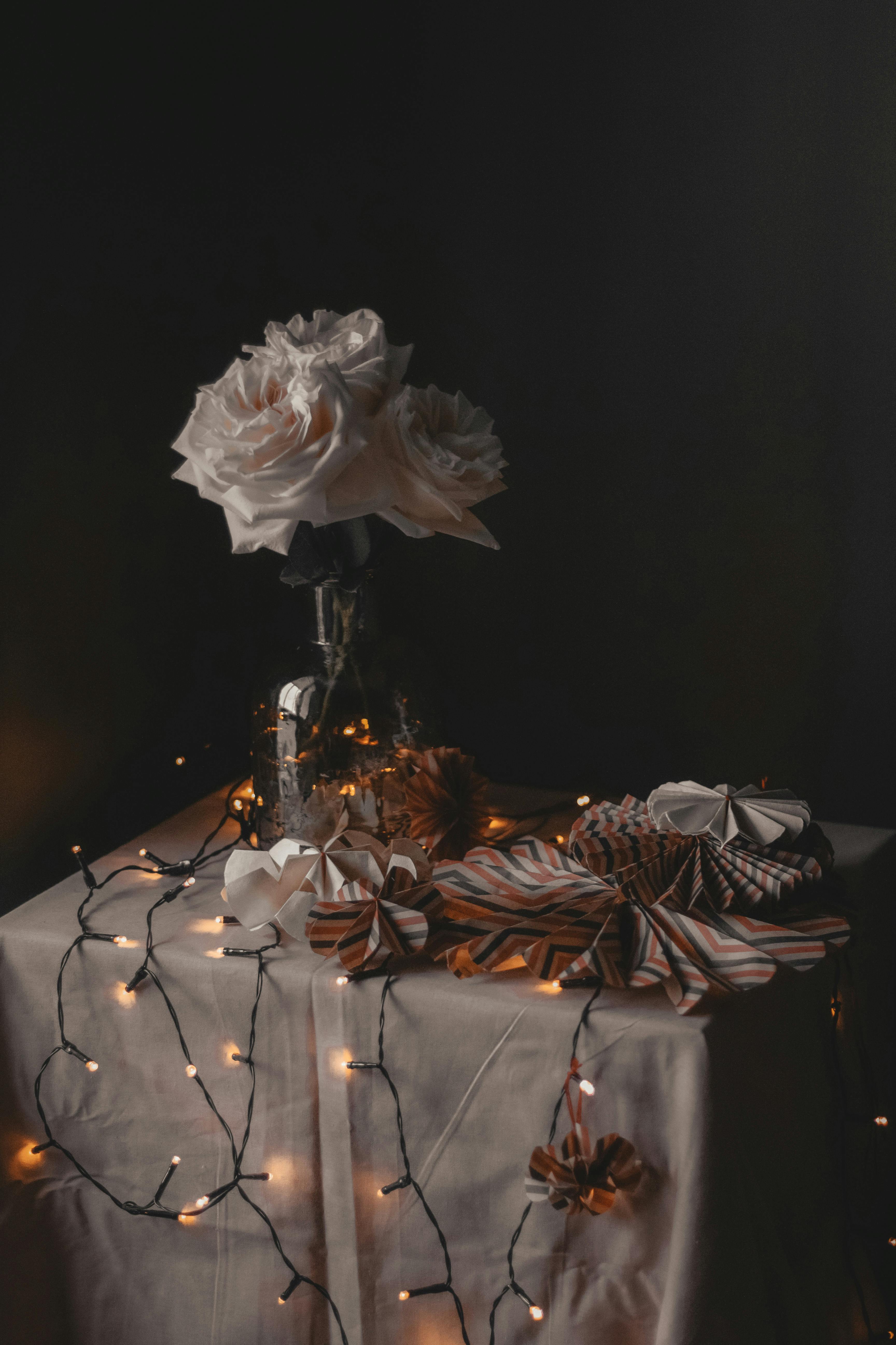 Composition of creative paper decorations placed on table with glowing garland and vase with blooming flowers in room on black background
