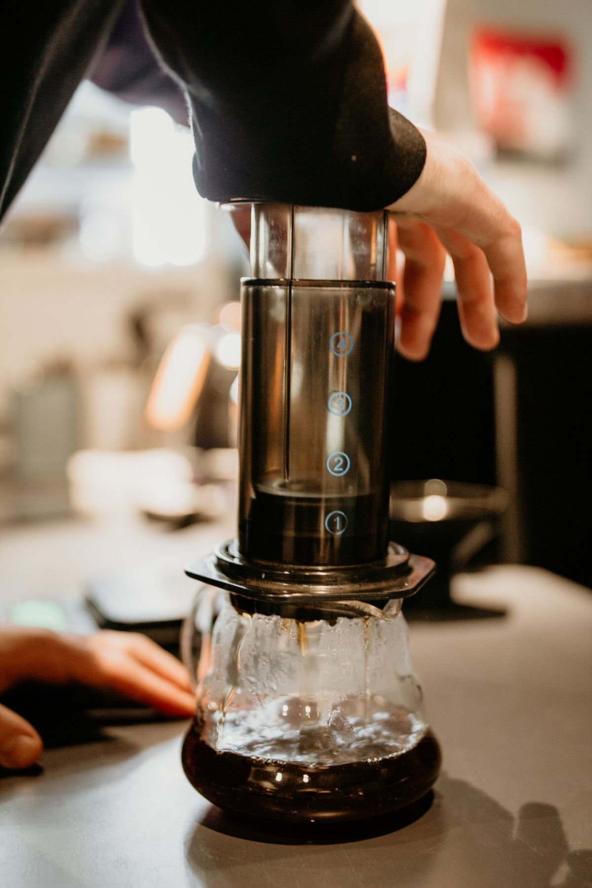 Barista apasă pistonul AeroPress peste o carafă de sticlă în timpul extracției cafelei