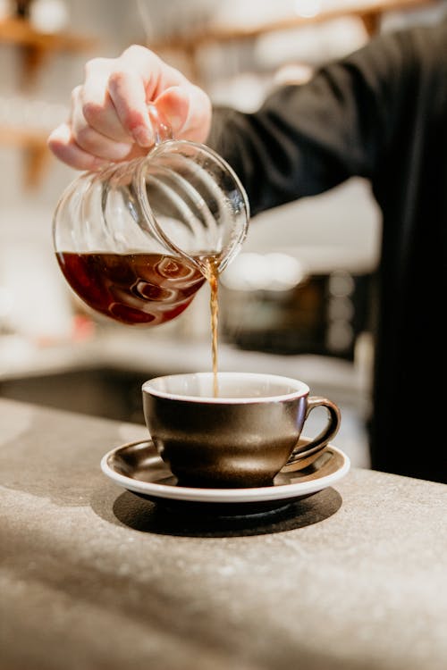 Unrecognizable professional barista pouring freshly brewed coffee from glass pot into cup while standing at counter on blurred background during work