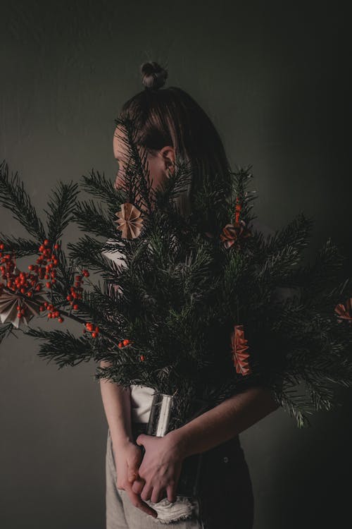Free Unrecognizable young female model standing with bunch of artificial red berry holly brunches and pine with paper holiday ornaments in glass vase against gray background Stock Photo