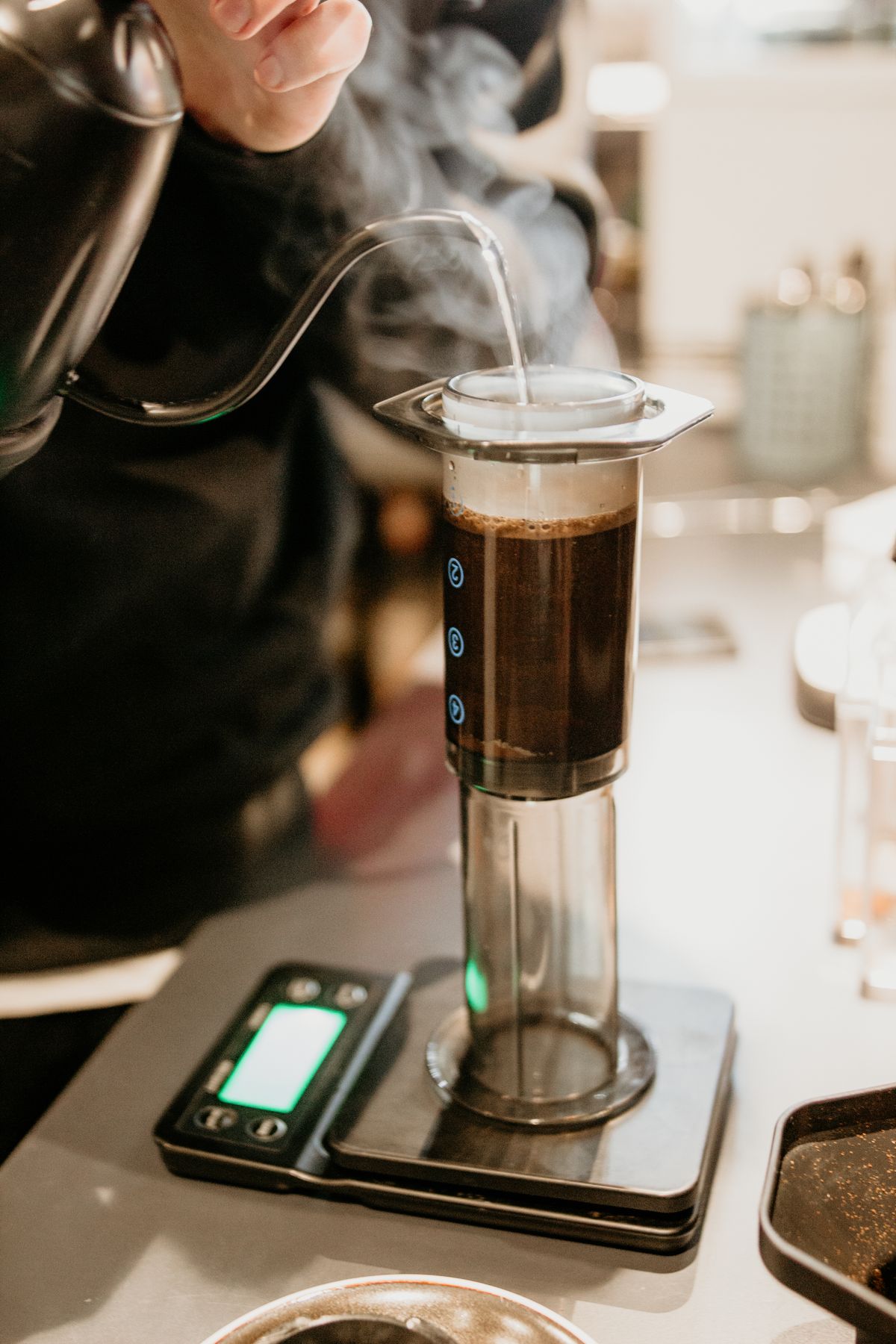 Barista toarnă apă fierbinte din ibric cu gât de gâscă într-un AeroPress pe cântar, cu abur ridicându-se