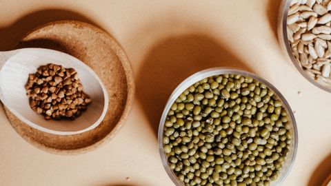 Top view of assorted grains and seeds including mung beans, buckwheat, and rice on a beige surface.