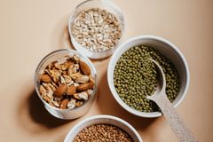 An overhead view of assorted nuts and grains in bowls with a spoon on a neutral background.