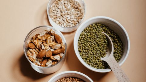 An overhead view of assorted nuts and grains in bowls with a spoon on a neutral background.