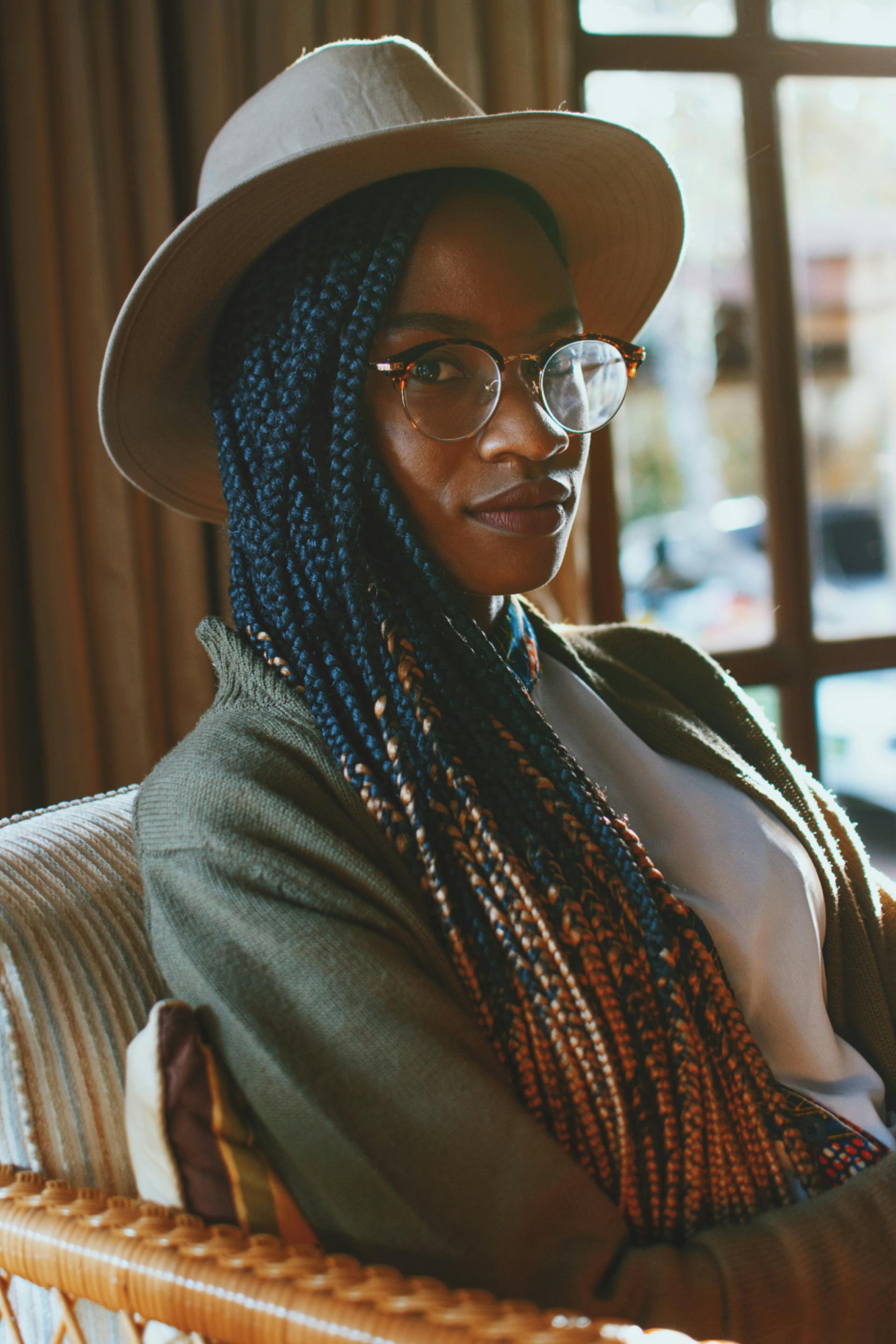 A fashionable woman with braided hair and glasses wearing a hat, sitting indoors by a window.