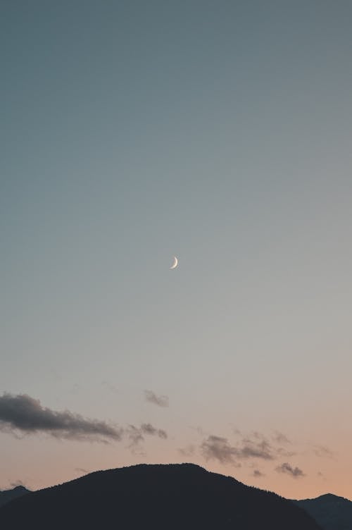 Small distant crescent moon shining on sundown sky with clouds above dark silhouette of mountain in evening time in nature