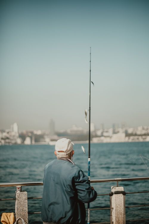 Free Elderly man enjoys tranquil fishing on a seaside pier at sunset with a distant city view. Stock Photo