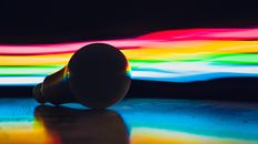 Single light bulb placed on dark surface against abstract background with multicolored rainbow lights in studio with black wall and dim light