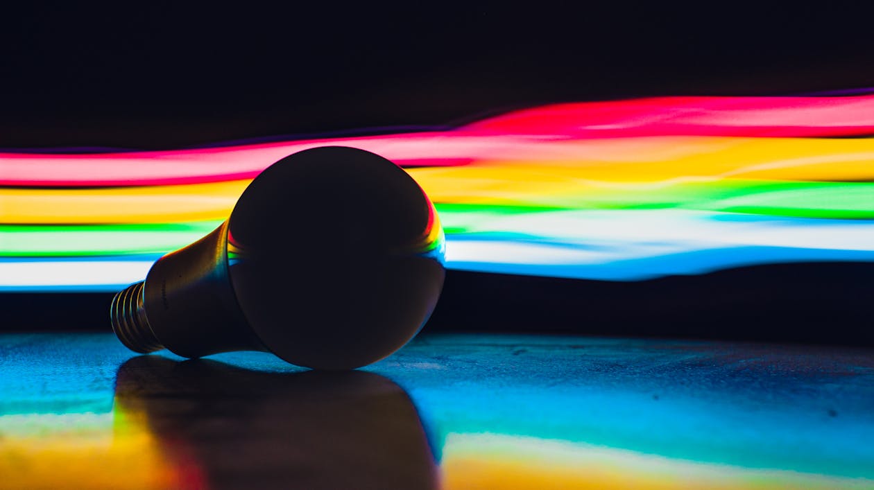 Single light bulb placed on dark surface against abstract background with multicolored rainbow lights in studio with black wall and dim light