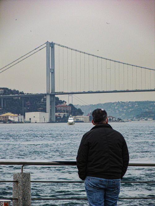 Free A man stands by the water in Istanbul, viewing the Bosphorus Bridge under an overcast sky. Stock Photo