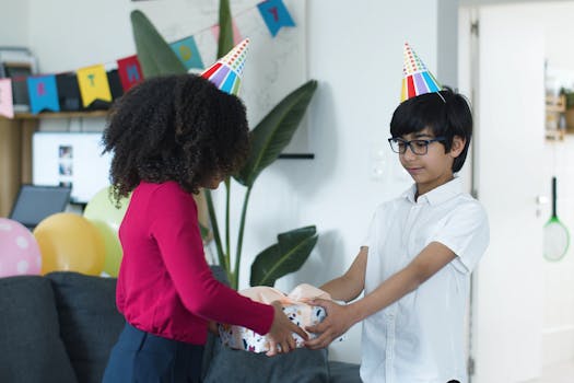 Two kids wearing party hats exchange a gift amidst colorful balloons in a cozy indoor setting.