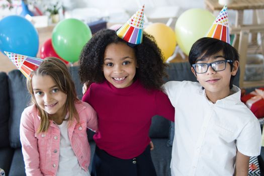 Three kids smiling and celebrating a birthday with colorful balloons and hats indoors.