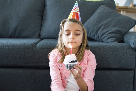 A young girl wearing a party hat holds a cupcake with a lit candle, celebrating her birthday indoors.