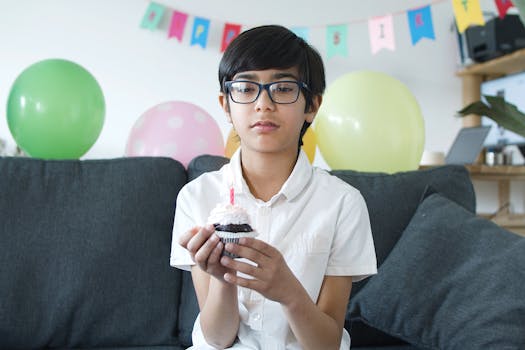 A young boy in eyeglasses holding a cupcake with a candle, celebrating his birthday indoors.
