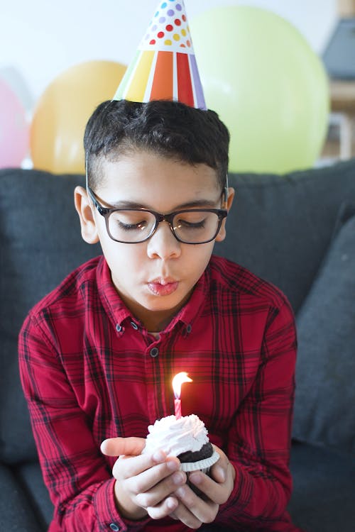 A Young Boy Wearing a Party Hat while Blowing His Cupcake