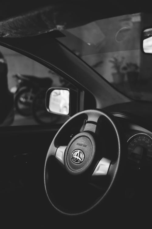 Black and white of steering wheel of modern automobile with speedometer and side mirror parked on street with transport in city