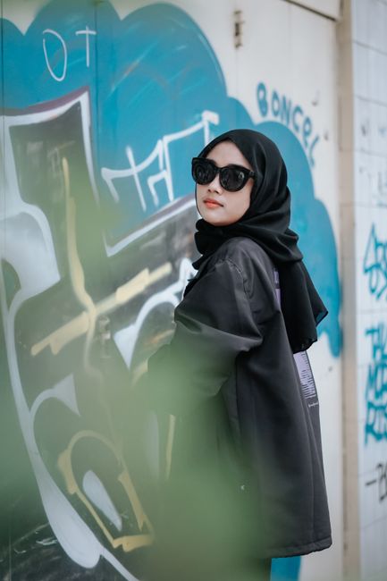 Elegant woman wearing a black hijab and sunglasses posing by a graffiti wall outdoors.