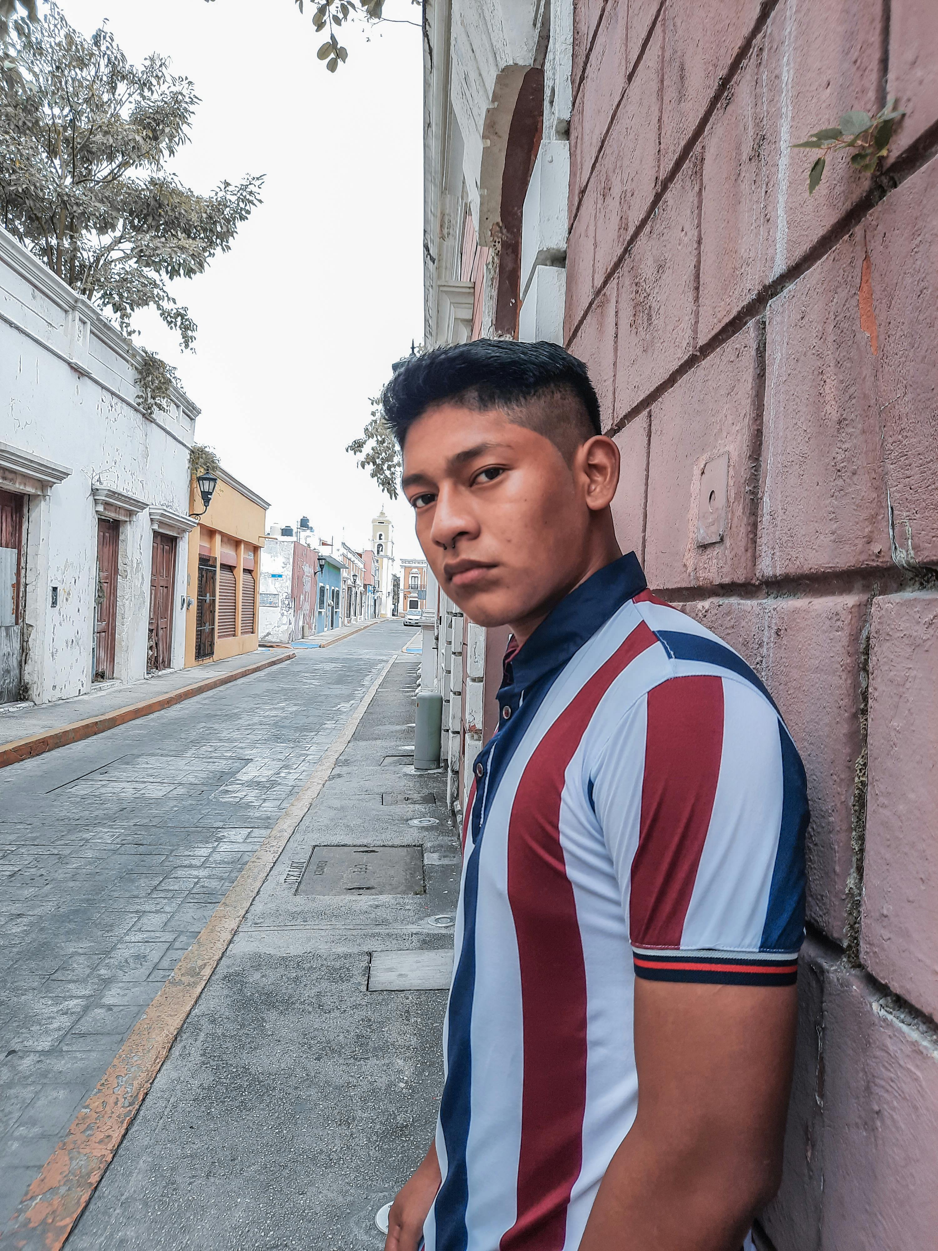 Portrait of a man in a striped shirt standing on a quiet urban street, leaning against a wall.