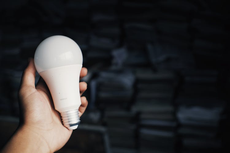 Person Holding Light Bulb Against Heap Of Books