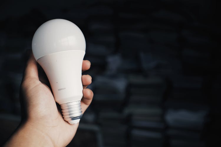 Person Reaching Out Hand With Light Bulb Against Books In Dark Room