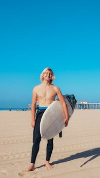 Smiling young surfer with surfboard on a sunny sandy beach during daytime.