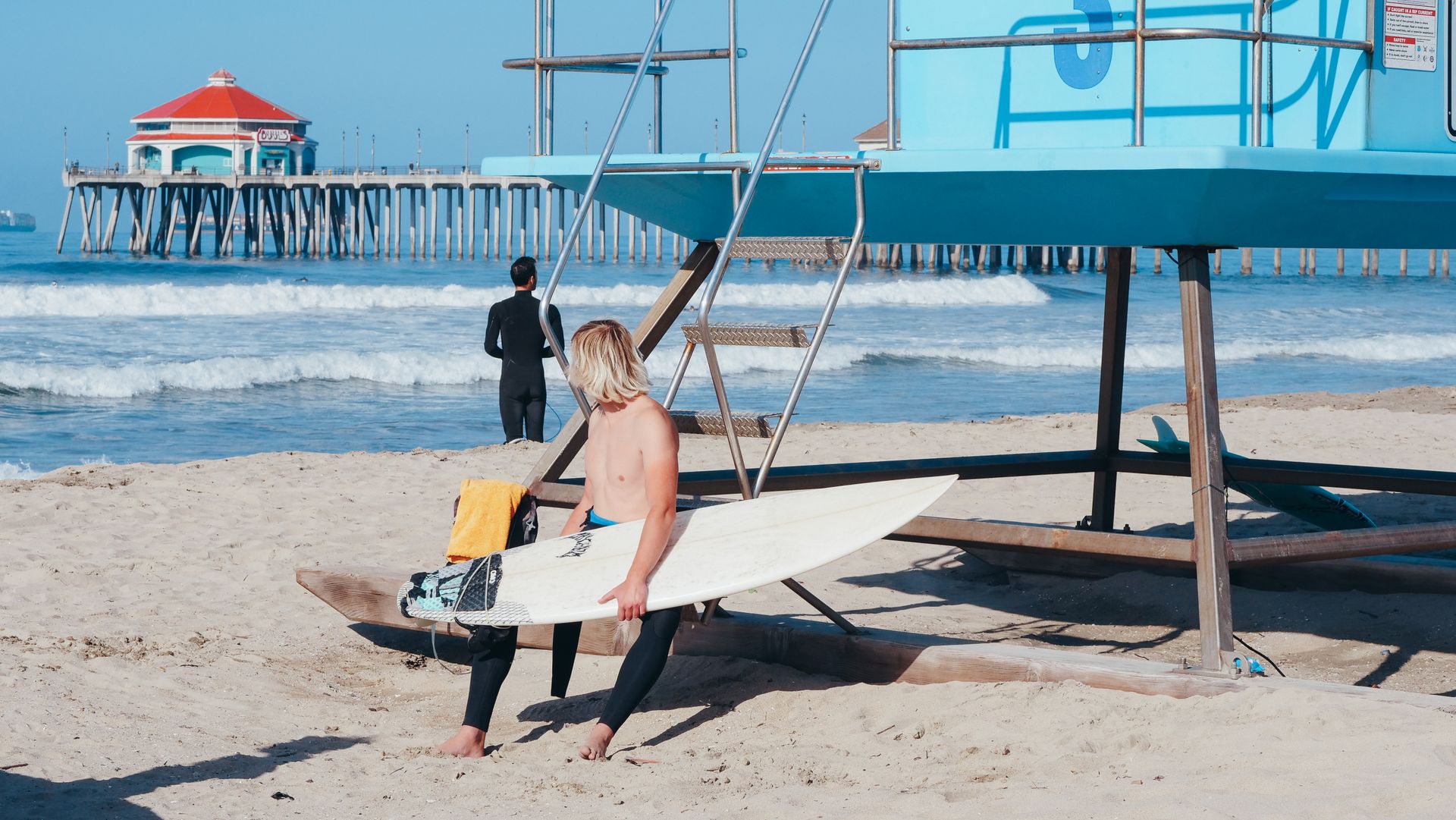 A surfer sits with a surfboard by a lifeguard tower at Huntington Beach pier.